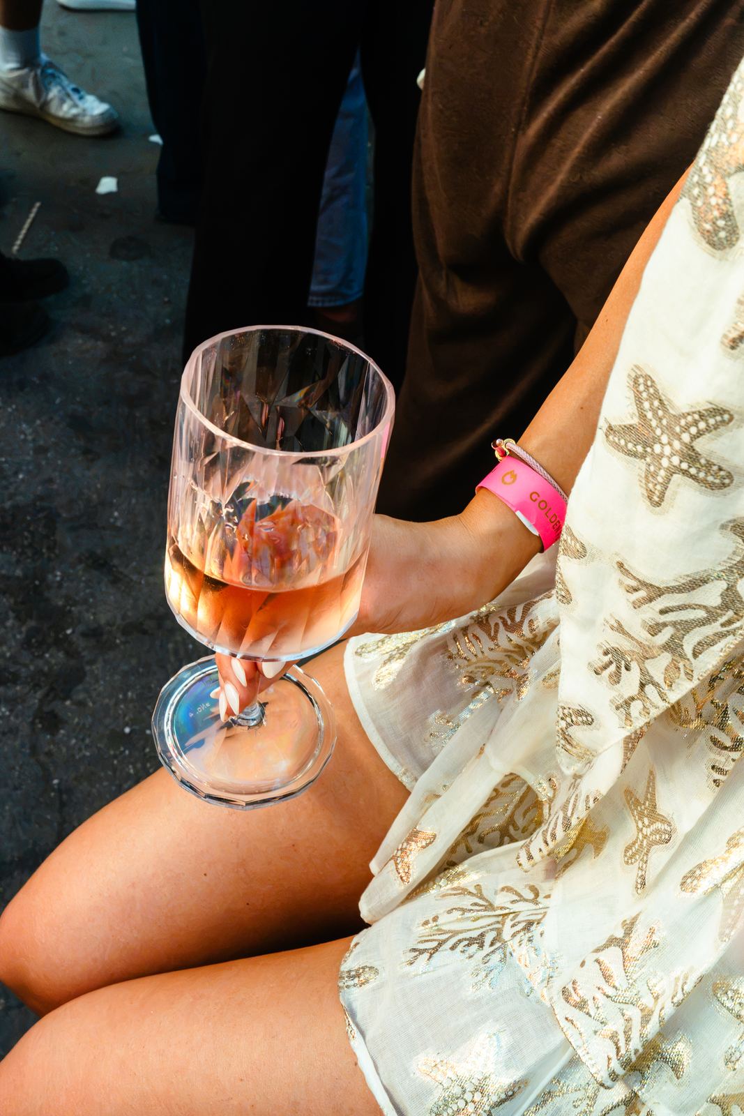 Hand hält ein Glas HEAVEN Rosé über einem Sommerkleid mit goldenen Sternen, dazu ein pinkes Event-Bändchen